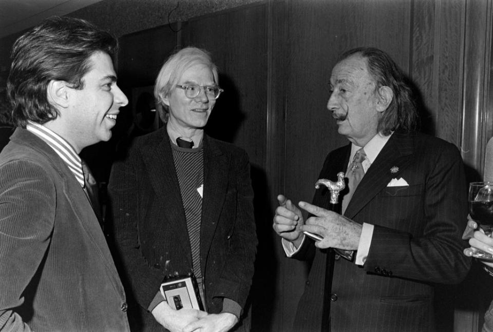 Three men appear conversing at an event: Robert Kardashian, in a suit; Andy Warhol, in casual attire with glasses; Salvador Dali, in a suit, holding a cane
