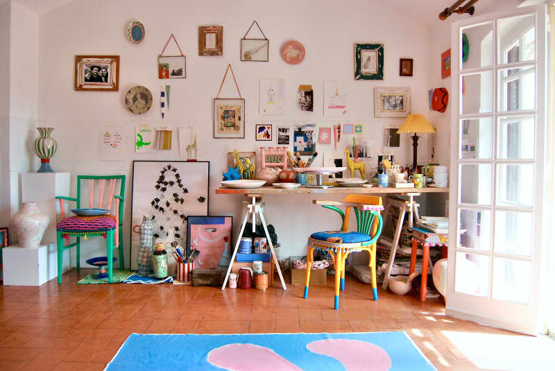 Art supplies on desk in white room with ceramics, gallery walls, and colorful chairs.