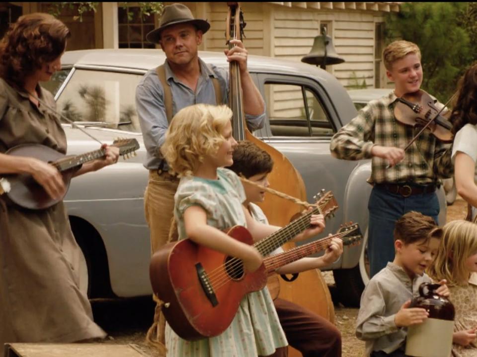 An actress playing a young Dolly Parton playing the guitar surrounded by other musicians outside.