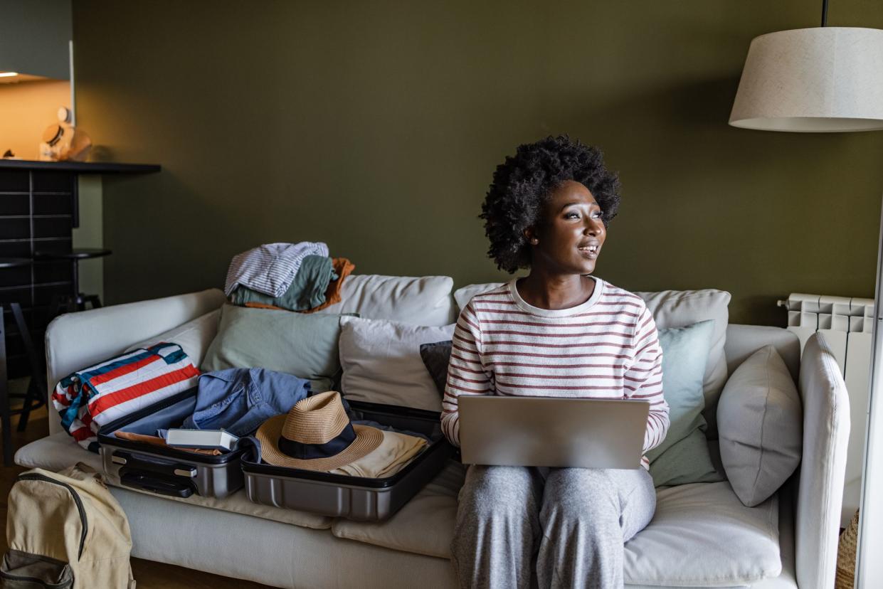 Young African American woman sitting on the couch in the living room and surfing the net on the laptop while packing her clothes for a trip