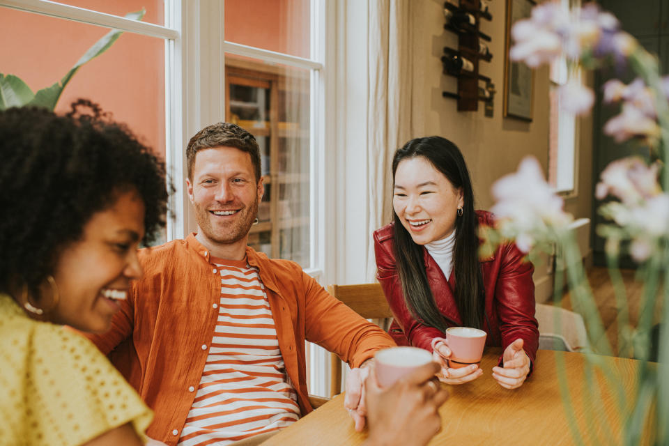 Three beautiful young people socialise at a table while enjoying hot drinks.  They are animated as they giggle together. One girl is in the foreground whilst focus remains on a smiling male and female.