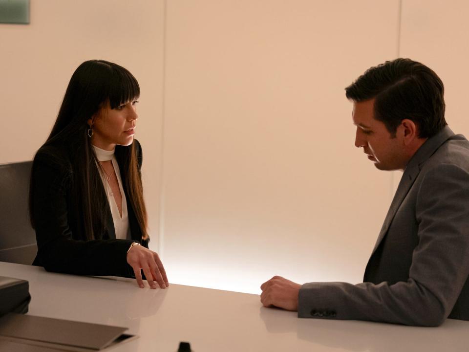 A white woman with long dark hair sitting across from a white man wearing a grey suit.