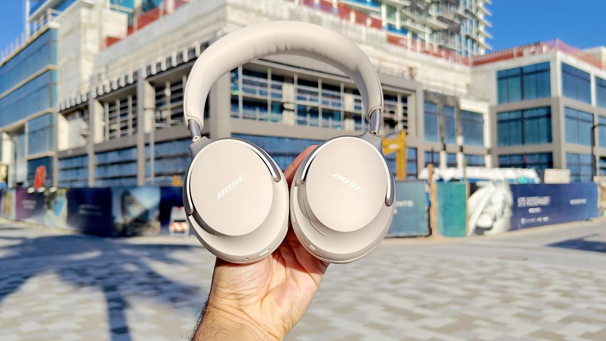  Bose QC Ultra Headphones listing image shown in smoke white held aloft in daylight on an urban street. 