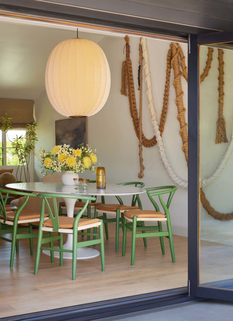Large sliding glass doors, green and woven modern dining chairs, white oval dining table, large paper mache lighting fixture, rope art installation