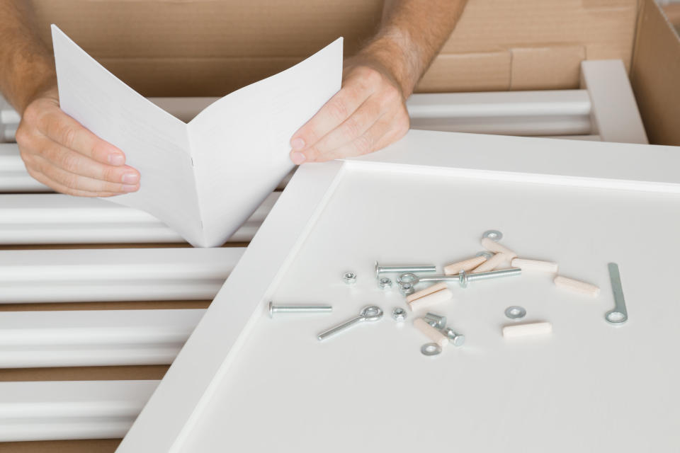 Hands holding instruction book. White wooden planks and screws for crib in cardboard box. Father preparation for future baby. Man assembling new furniture. Close up.