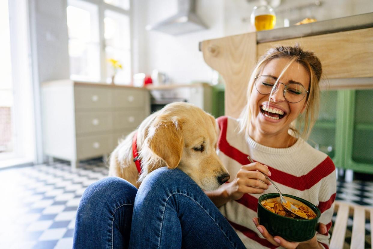 woman sitting on kitchen floor laughing and eating food next to her dog