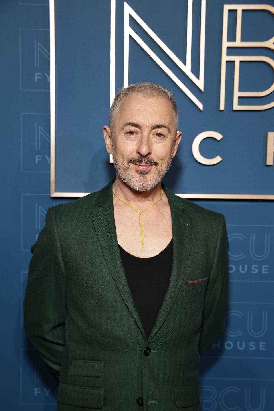 Alan Cumming in a green blazer and black shirt, posing in front of a backdrop at an event