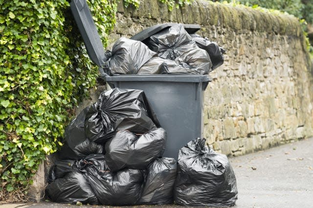 A full wheelie bin with rubbish bags overflowing onto the pavement.