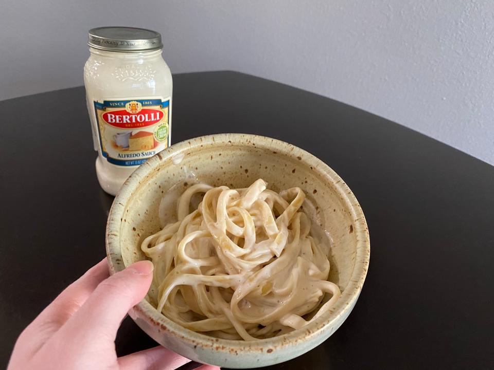 The writer holds a bowl of Alfredo with a jar of Bertolli Alfredo sauce in background
