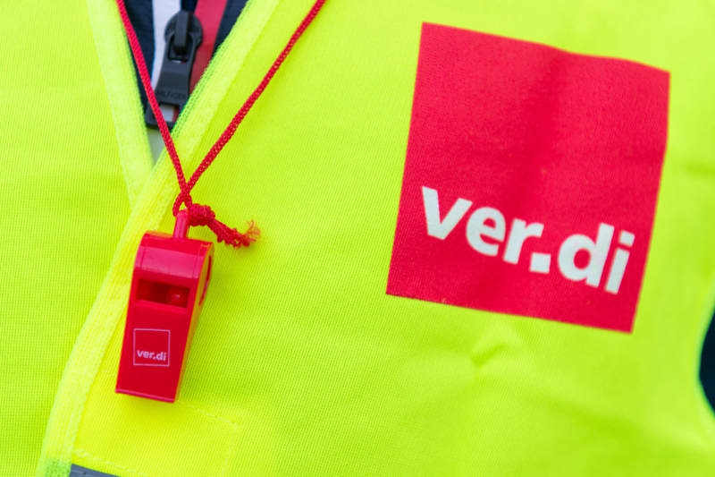 A person wears a high-visibility vest with the Verdi logo and a red whistle. Christophe Gateau/dpa