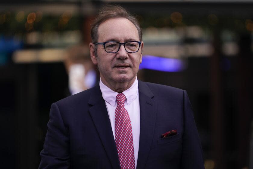 Kevin Spacey wearing glasses and in a dark suit, pink dress shirt and red tie standing against a blurred background