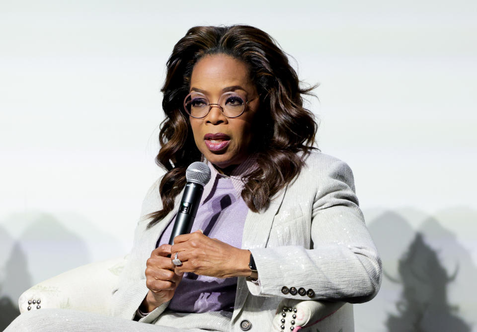 Oprah Winfrey is seated and holding a microphone, dressed in a stylish suit, speaking at an event