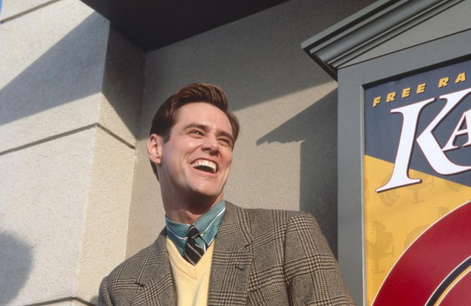 Jim Carrey as character Truman Burbank smiling, wearing a suit and patterned tie, beside a "Kaiser Chicken" poster