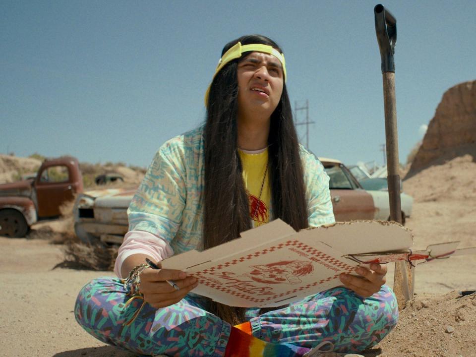 A young man with long hair sits on the ground in a sandy car lot.