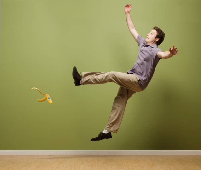 A man in a casual shirt and pants slips on a banana peel mid-air against a plain background