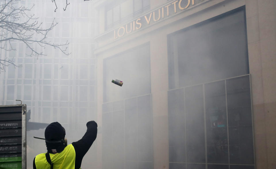 A protester throws a device on the Louis Vuitton luxury shop on the Champs Elysees avenue, during a yellow vests demonstration Saturday, March 16, 2019 in Paris. Paris police say more than 100 people have been arrested amid rioting in the French capital by yellow vest protesters and clashes with police. They set life-threatening fires, smashed up luxury stores and clashed with police firing tear gas and water cannon (AP Photo/Christophe Ena)