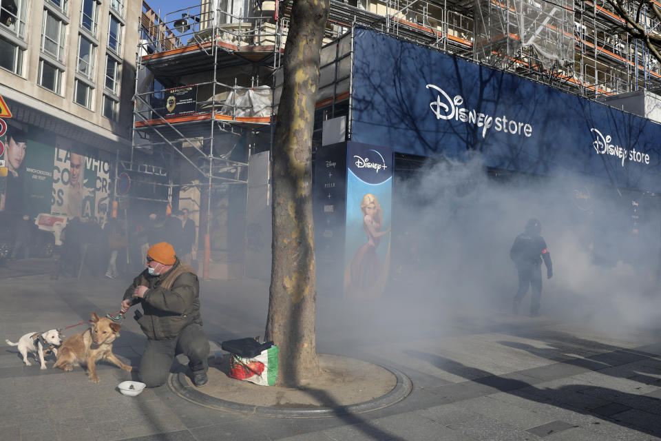 A man holds his dogs while police officer walks though tear gas during a protest on the Champs-Elysees avenue, Saturday, Feb.12, 2022 in Paris. Paris police intercepted at least 500 vehicles attempting to enter the French capital in defiance of a police order to take part in protests against virus restrictions inspired by the Canada's horn-honking "Freedom Convoy." . (AP Photo/Adrienne Surprenant)