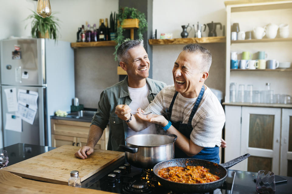 Two smiling men in a kitchen, cooking together. One is holding a tasting spoon and laughing, while the other watches him affectionately