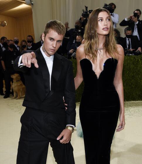 Justin Bieber in a baggy black suit pointing at the camera while standing next to Hailey Bieber in a black strapless gown