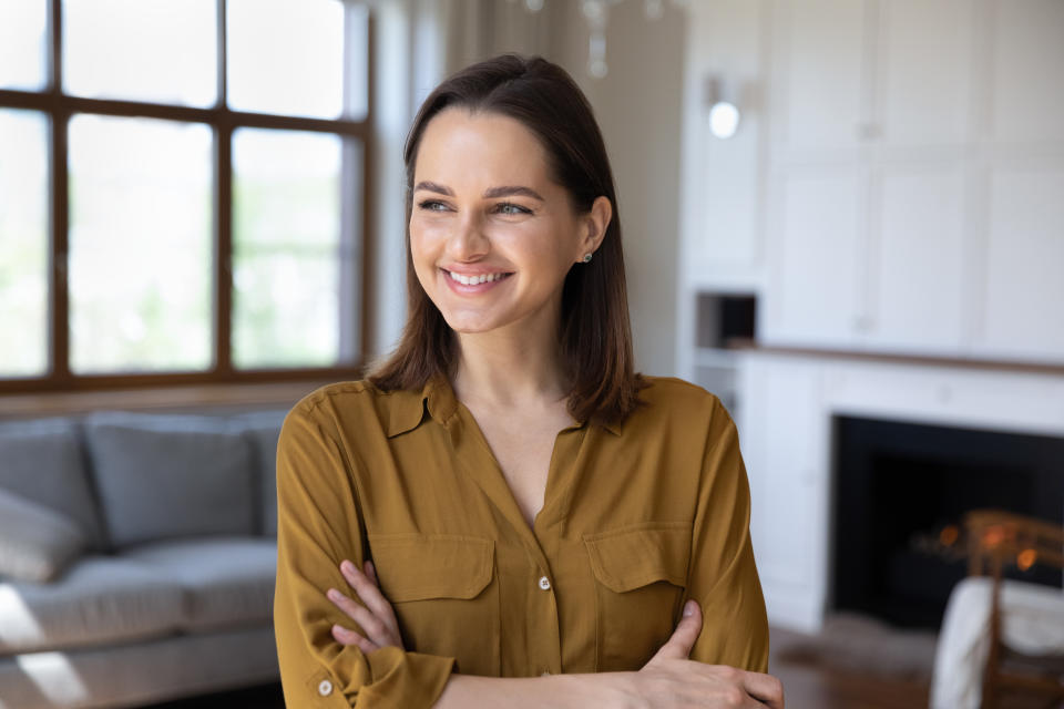 Happy confident house tenant woman enjoying being at new home, standing in living room interior with folded arms, looking away with thoughtful face, smiling at good thoughts, plans, future vision