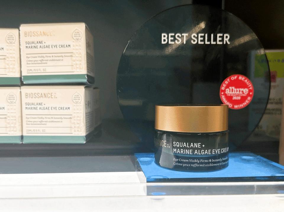 A dark-green container of Biossance squalane + marine algae eye cream on a shelf in front of cream-colored boxes at Sephora