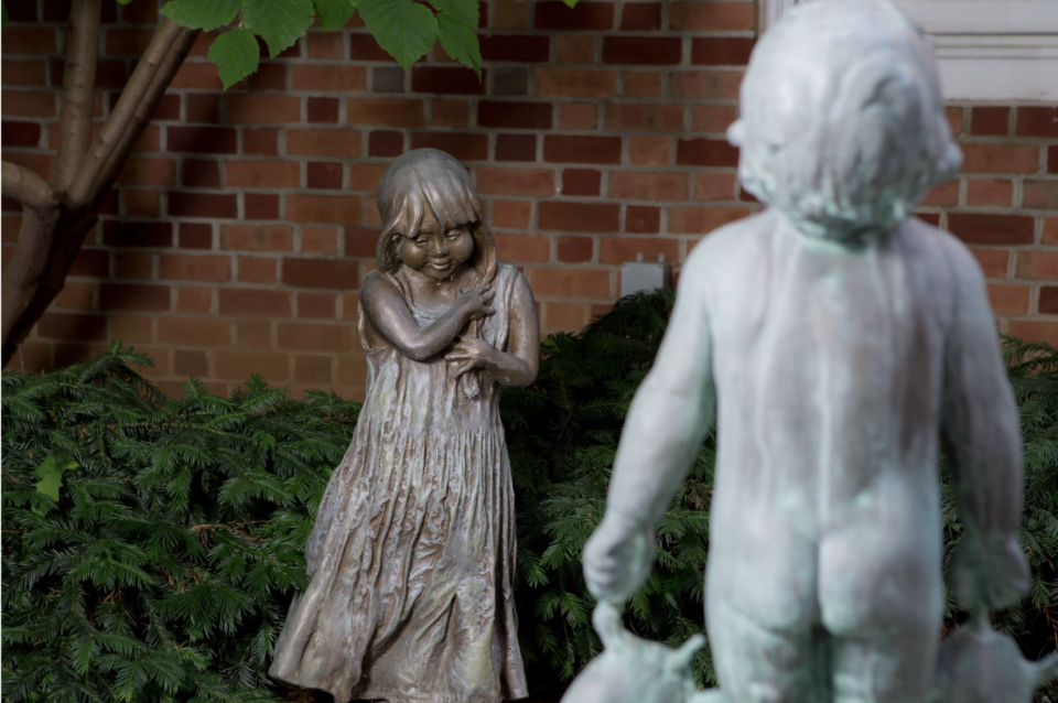 Lorann Jacobs’ sculpture of a little girl holding flowers shares space in Martin Library’s Sensory Garden with the bronze piece that inspired her as a child, the “Turtle Baby.” Today, the two art pieces stand side-by-side in the library’s atrium until renovation work on the library’s children’s wing and the nearby garden is finished. When re-opened in 2022, the garden will be available for members of the public to walk through where they, too, can share space with the two figures.