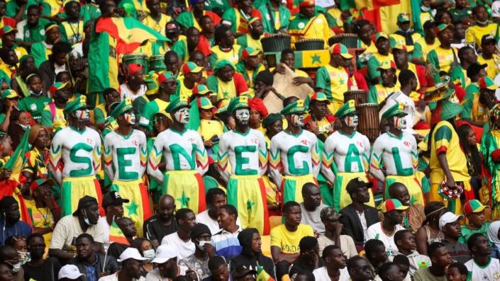 Senegalese people with face paint on and clothing in the Senegalese flag colours at a stadium. They have Senegal written out on their chests. It is quite a large and packed crowd.