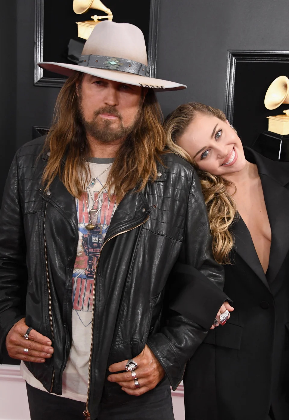 Billy Ray Cyrus wearing a leather jacket and hat, posing on the red carpet with Miley Cyrus, who is resting her head on his shoulder and smiling