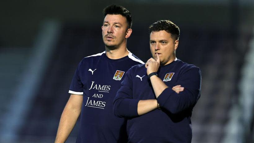 Northampton Town joint managers Liam Williams and Josh Oldfield both with short dark hair and wearing blue shirts standing on a football pitch