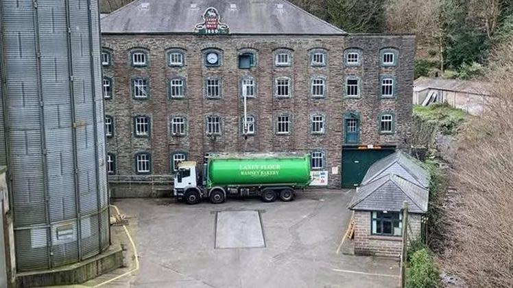 A small office block set in front of a mill building with a green flour truck parked in front of it and a flour silo to the left hand side