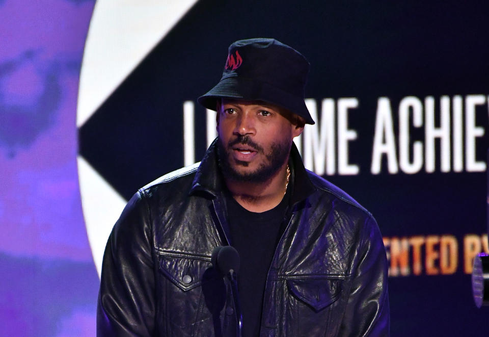 Marlon Wayans speaks on stage wearing a black leather jacket and a black bucket hat at an event