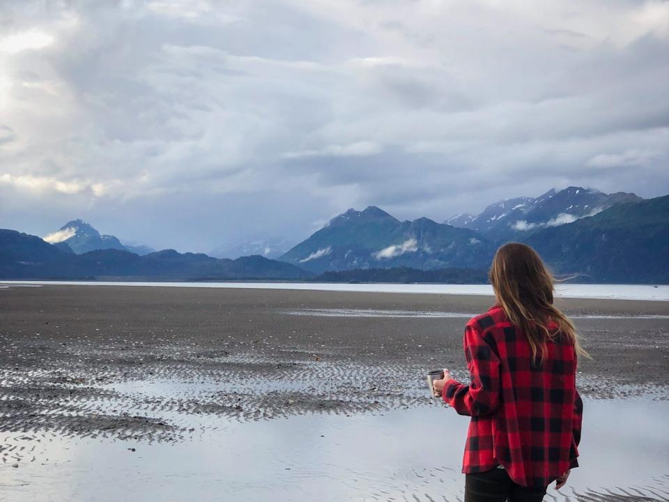 Emily walking toward wet sand, with mountains in the distance. Her back is to the camera and she's wearing a red-and-black flannel, black pants, and red boots.