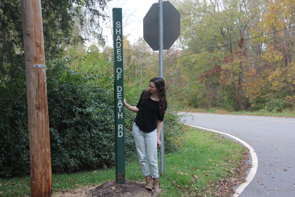 A woman stands next to a road sign reading "Shades of Death Road."