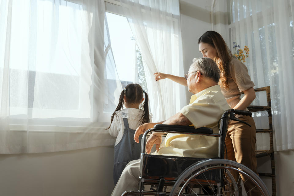 The elder in a wheelchair plays with his family lovingly in the house, the morning sun shines through the glass.