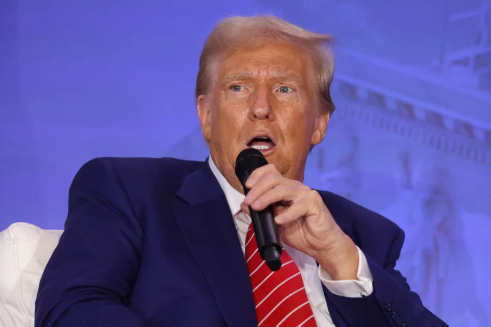 Donald Trump is speaking into a microphone while seated, wearing a suit and striped tie