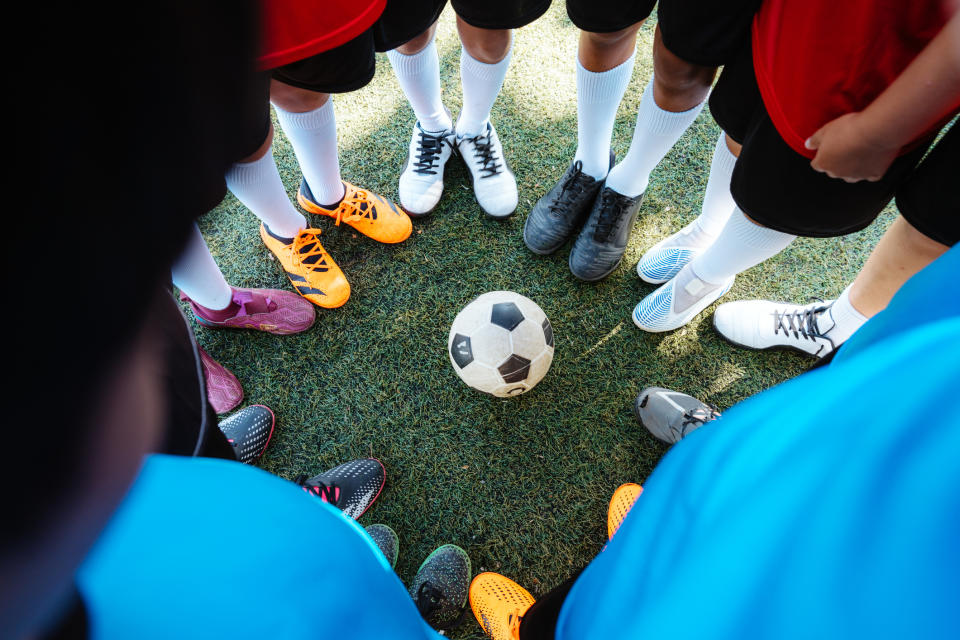 A group of unidentified people in soccer uniforms standing in a circle, their feet surrounding a soccer ball on a grass field