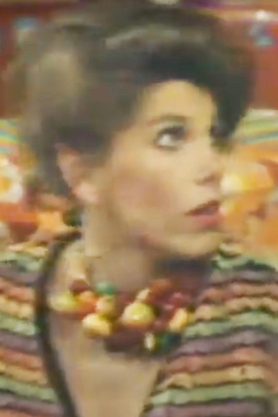 Christine Baranski wearing a colorful striped sweater and a chunky necklace looks startled on a brightly decorated set