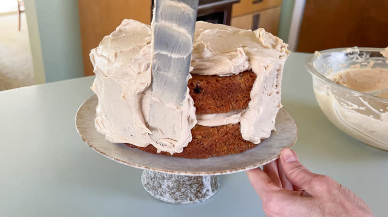 Spreading cinnamon-cashew frosting on vegan carrot cake with spatula on serving platter