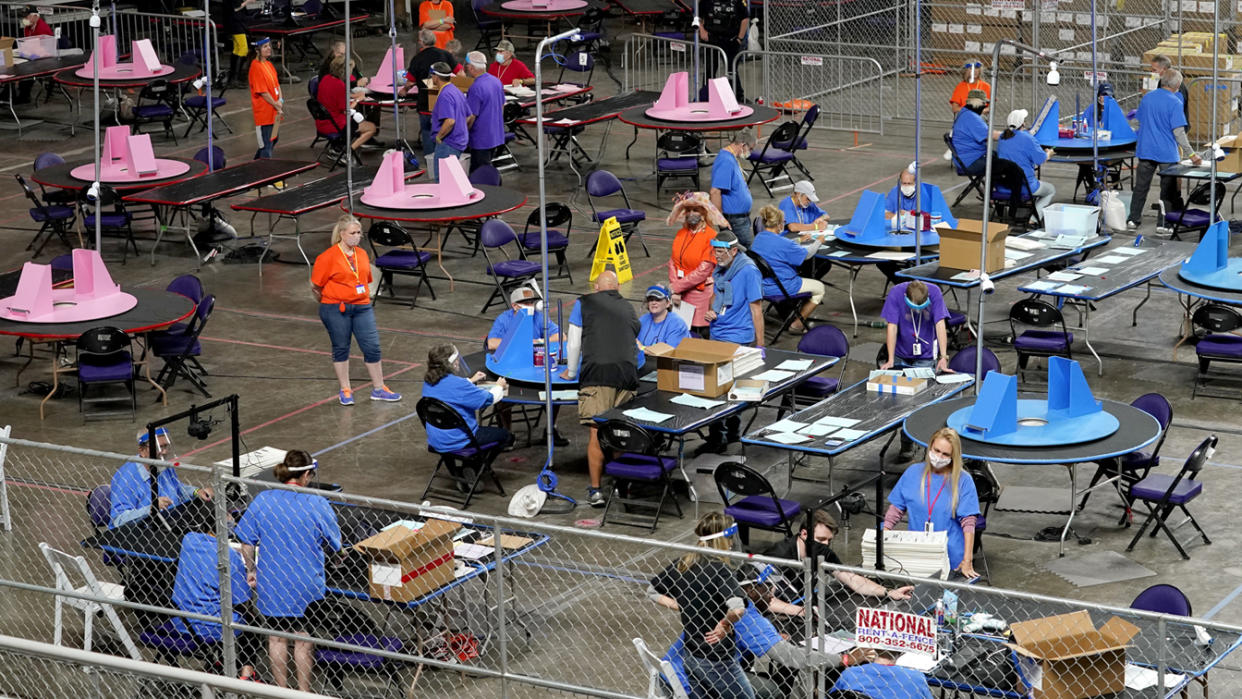 Several dozen people wearing red, blue or purple shirts with lanyards around their necks stand or sit at round tables with blue or pink vote-counting materials on them in a large room penned in by chain-link fencing. 