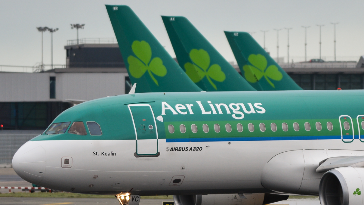 Green and white Plane with Aer Lingus written across it with three plane tail wings in the background that each have the Aer Lingus shamrock logo on them