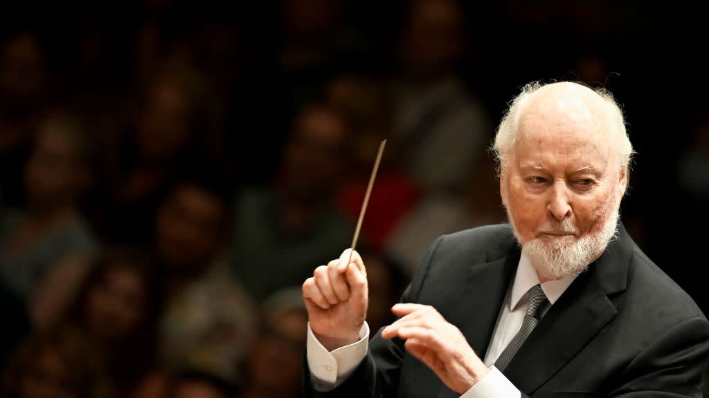 john williams holds a baton while waving his arms, he looks to the right and wears a black suit jacket, silver tie and white collared shirt