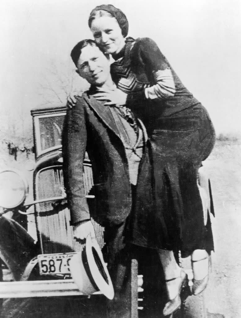 A couple from the early 20th century poses in front of a vintage car; the woman stands on the car's side, holding the man's shoulders