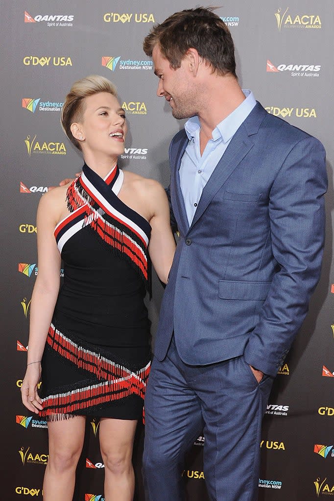 Scarlett Johansson and Chris Hemsworth pose together at an event. Scarlett wears a stylish black and red dress; Chris dons a sharp blue suit