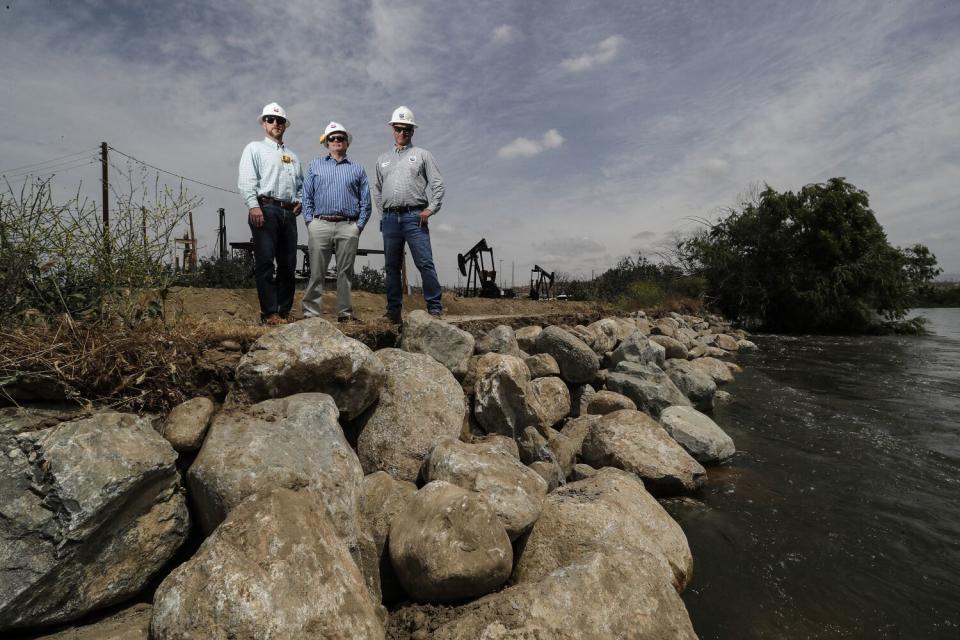 Three men in hardhats stand on a riverbank.