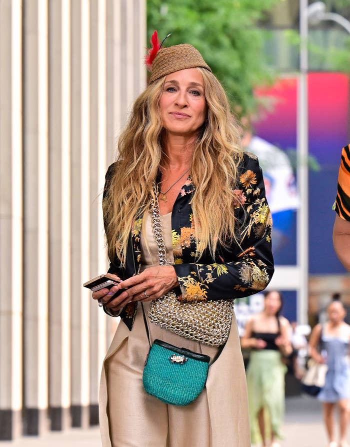 Sarah Jessica Parker wears a stylish outfit featuring a floral jacket, beige hat with a red accent, beige dress, and a blue purse while walking in an urban setting