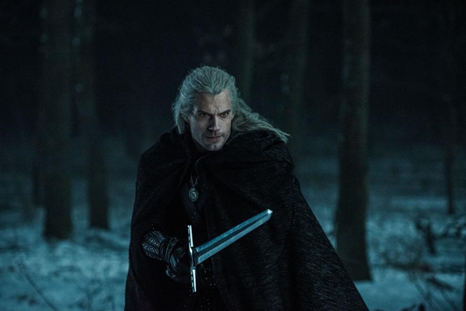 Geralt the witcher wearing a cloak with his sword drawn in the forest