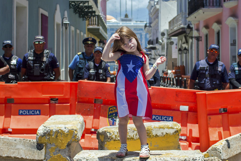 A girl wearing a dress featuring the Puerto Rican flag stands in front of police blocking the road leading to the La Fortaleza governor's mansion in San Juan, Puerto Rico, Thursday, July 18, 2019. Protesters are demanding Gov. Ricardo Rossello resign after the leak of online chats that show him making misogynistic slurs and mocking his constituents. (AP Photo/Dennis M. Rivera Pichardo)