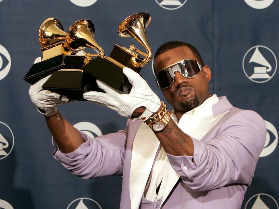 Kanye West holding multiple Grammy awards while wearing sunglasses, a white shirt, a lavender jacket, and white gloves, posing on a red carpet