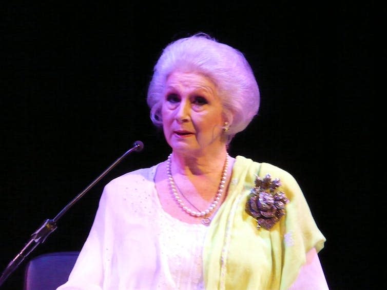 April Ashley, an elderly woman with a white pouf hairstyle, speaking onstage.