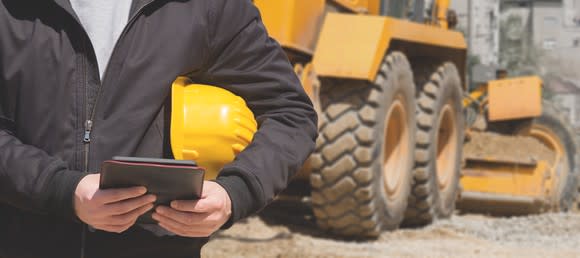 A construction worker with a tablet standing in front of heavy equipment.